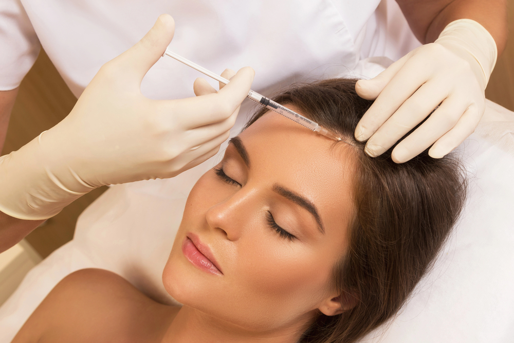 Medical professional in latex gloves administering a forehead injection to a relaxed woman lying on a treatment table.